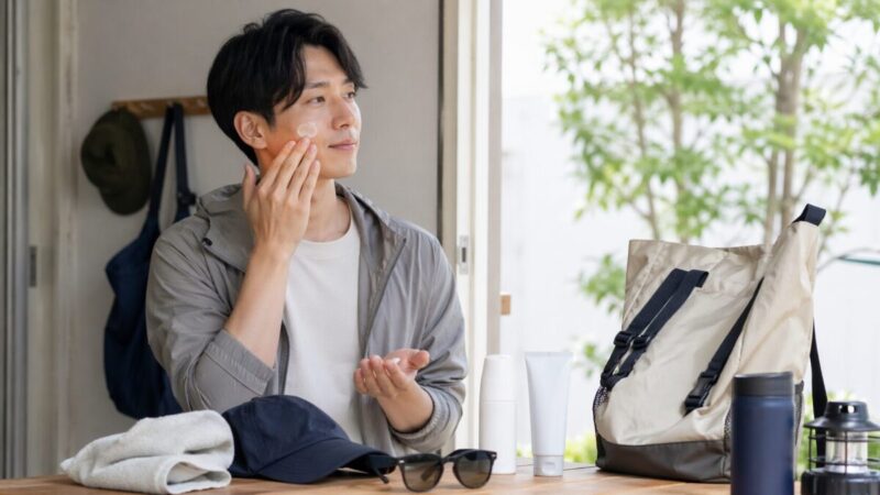 外出前に頬へ日焼け止めをなじませる男性とアウトドア小物のイメージ