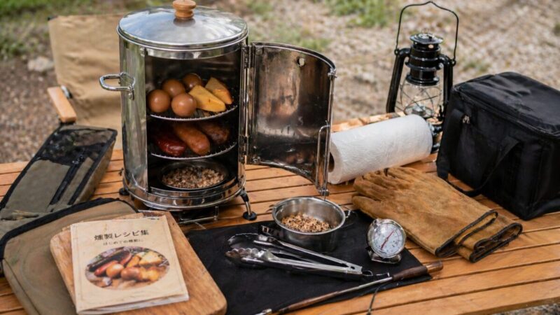 キャンプテーブルに並べた燻製器と温度計、グローブ、収納用品を確認するレビュー用の道具一式