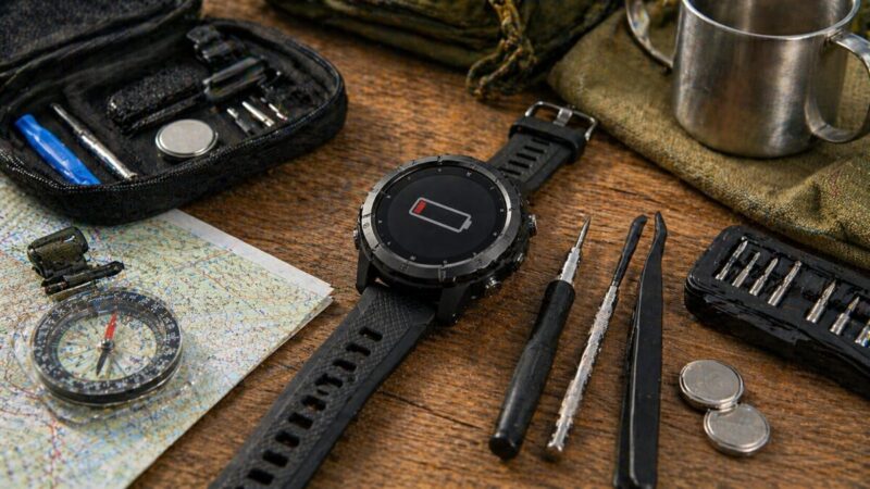 アウトドア用スマートウォッチと工具、交換用電池、登山用品を並べた修理費用と買い替え比較の写真。