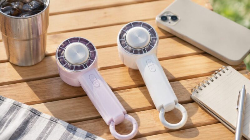 屋外テーブルに冷却プレート付きハンディファンとスマートフォン、メモ帳、飲み物を並べた様子