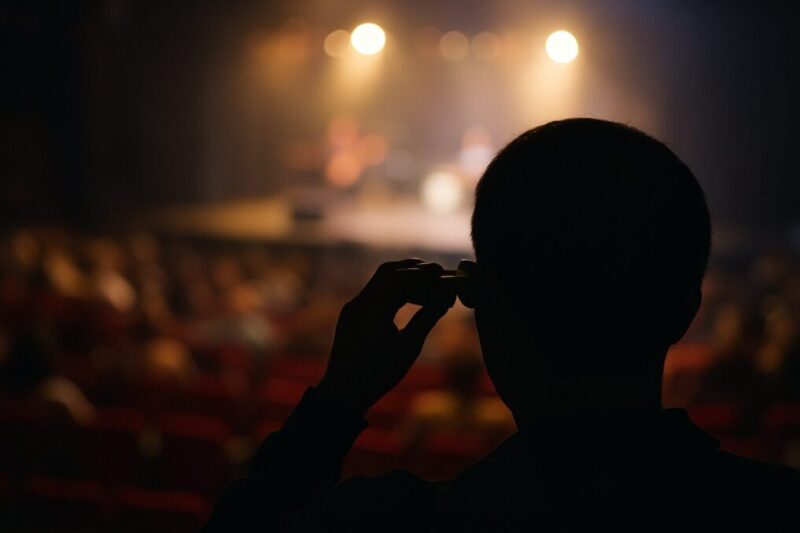 客席から双眼鏡を構えてステージ方向を見つめる後ろ姿をシルエットで写し、ステージの光がぼけて広がる距離感を表したイメージ