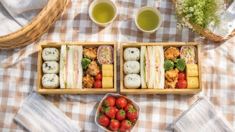 おにぎりやサンドイッチ、唐揚げ、卵焼き、いちごを二人分並べたピクニック弁当