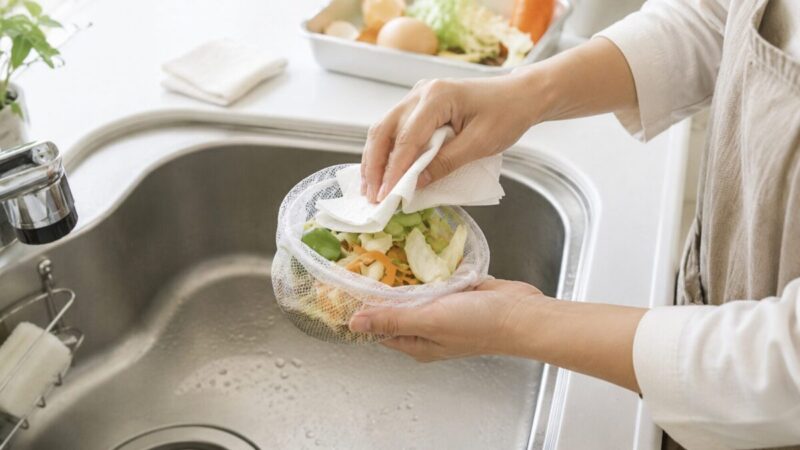 キッチンのシンクで、野菜くずの水分をペーパーで拭き取りながら水切りネットにまとめている様子。