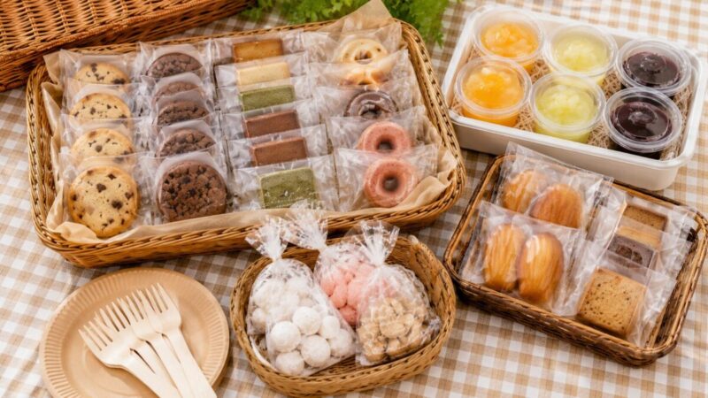 個包装のクッキー、焼き菓子、ミニドーナツ、カップゼリーをラタンバスケットに並べ、紙皿と木製フォークを添えた持ち寄り用スイーツ。