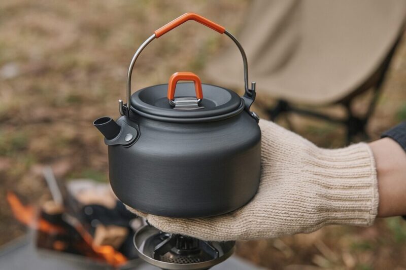 外の焚き火台の前で、日本人が耐熱グローブで黒い小型ケトルのベイルハンドルを握り、短い注ぎ口と低い背が分かる角度で持ち上げている