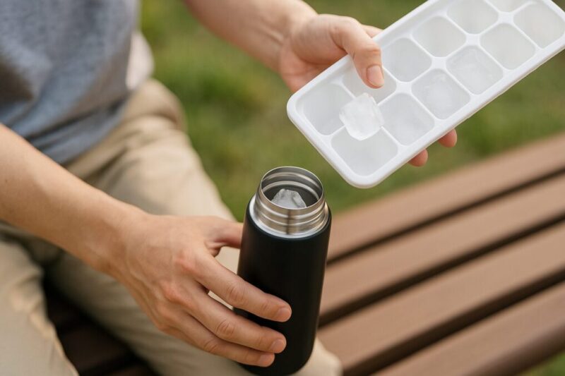 屋外ベンチで、広口のステンレスボトルに製氷トレーから氷を投入する日本人の手元